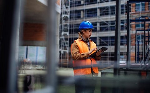 An employee wearing a high-visibility jacket and blue helmet, looking at a laptop.