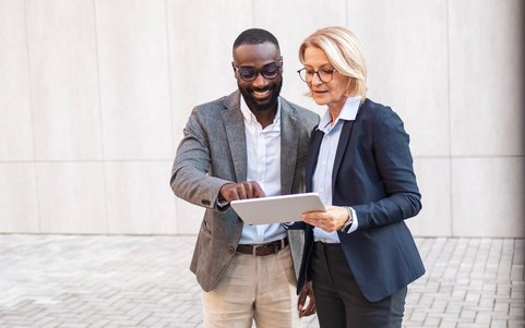 Two business professionals standing outside a building, reviewing content on a tablet together.
