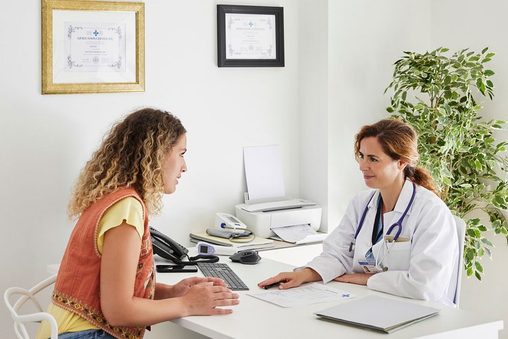 Doctor in a white coat consulting with a seated patient across a desk in a bright medical office.