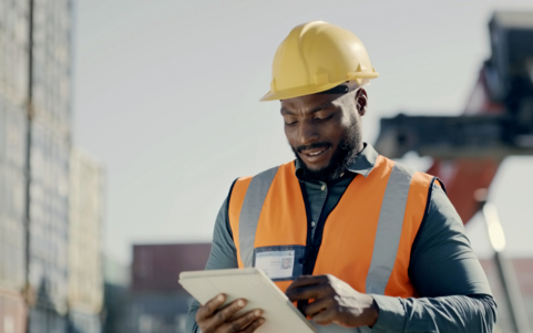 Worker in a yellow hard hat and orange high-visibility vest uses a tablet outdoors at an industrial site