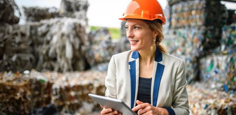 Woman in an orange hard hat and blazer holding a tablet at a recycling facility with stacked waste bales.