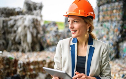 Woman in an orange hard hat and blazer holding a tablet at a recycling facility with stacked waste bales.
