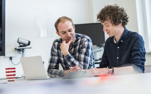 Two colleagues sitting together at a desk, looking at a laptop screen in a bright office.