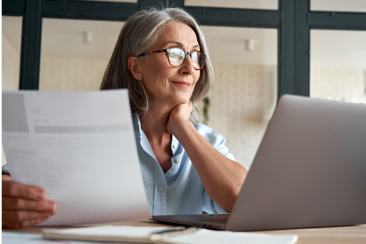 Woman with glasses reviews a document next to her laptop in a bright office