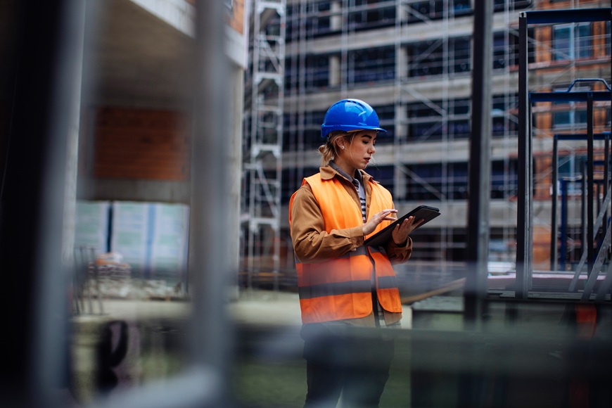 An employee wearing a high-visibility jacket and blue helmet, looking at a laptop.